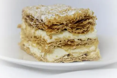 Deliciosa porción de costrada de Soria, con capas crujientes de hojaldre y crema suave, espolvoreada con azúcar glas.