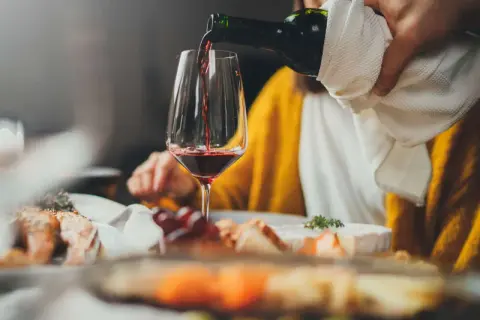 Rödvinet hälls upp i ett glas vid en middagsbjudning. En person i gul tröja sitter bakom. Frågan om hur mycket vin per person är relevant vid sådana tillfällen.