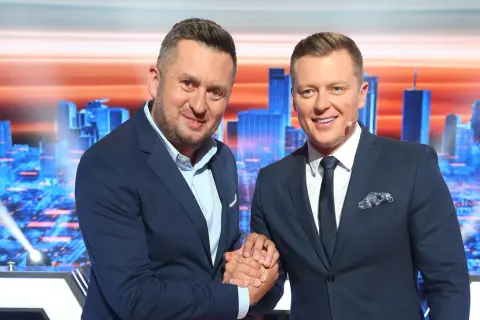 Dwóch mężczyzn w garniturach ściska sobie dłonie na tle scenografii z miejskim krajobrazem, gdzie nagrywane jest koło fortuny.