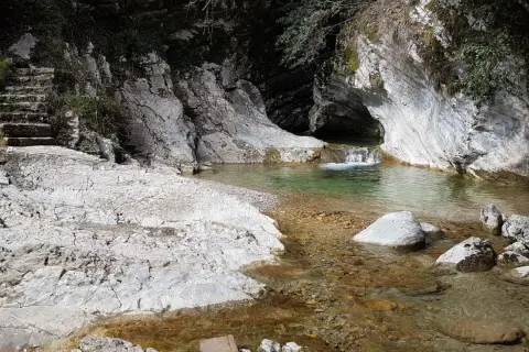 Sentiero di pietra conduce a un ruscello cristallino con piccole cascate di piovere, incastonato tra rocce bianche e vegetazione.