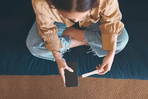 Kobieta siedzi ze skrzyżowanymi nogami, trzymając w dłoniach telefon i test ciążowy.