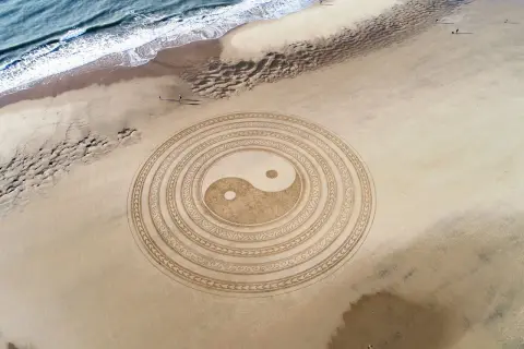 Símbolo Yin Yang en la arena, representando la energía masculina y femenina en equilibrio. El mar y la playa completan la escena.