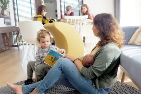 Mama karmi piersią niemowlę, podczas gdy starsze dziecko czyta książkę. To piękny przykład karmienia mieszanego w rodzinnej atmosferze.