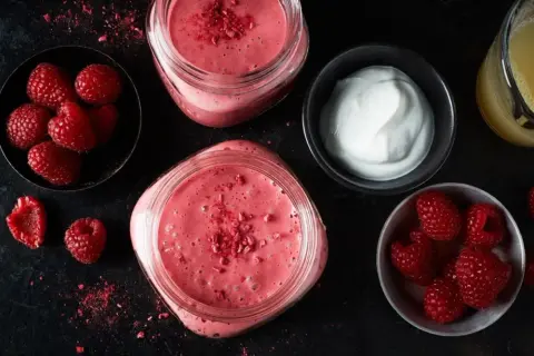 Två glas med en rosa, nyttig smoothie toppad med frystorkade hallon. Bredvid finns skålar med färska hallon och yoghurt, samt ett glas juice.