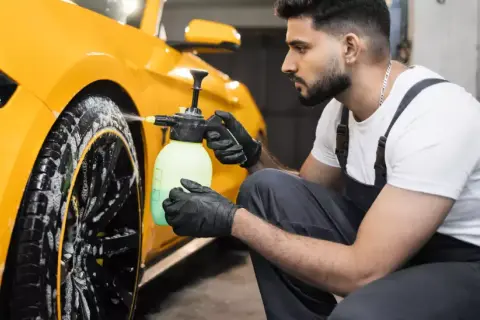 Mann mit Handschuhen sprüht Reinigungsmittel auf gelbe Felgen eines Autos.