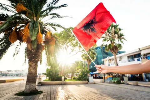 Dłoń trzyma flagę Albanii na tle palmy i słońca. Czy Albania jest w Schengen? To pytanie nurtuje wielu podróżnych.