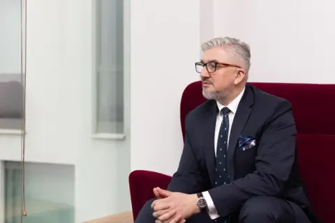 Mężczyzna w garniturze i okularach siedzi na czerwonym fotelu, patrząc w bok.