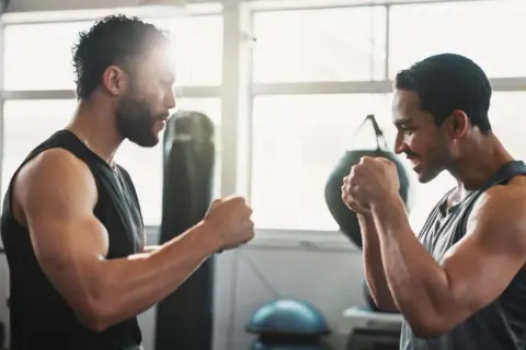 Two men in a gym, focused on their weight training for boxers. Punching bags and gym equipment are visible in the background.