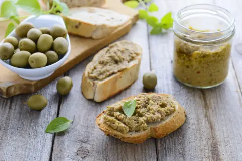 Bruschette con crema di olive, un vasetto di crema di olive e olive verdi su un tagliere di legno.