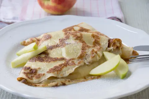 Zwei goldbraune Pfannkuchen mit Apfelmus gefüllt, garniert mit Apfelspalten und Puderzucker.
