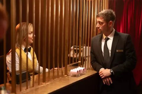 En kvinna i gul väst och fluga samt en man i kostym möts bakom en disk. En scen från en ny brittisk serie på SVT.