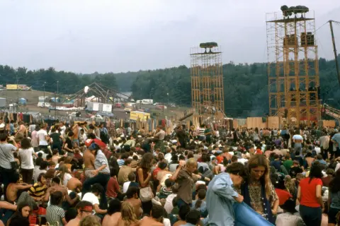 Tłum ludzi na festiwalu, gdzie jest Woodstock? Widać scenę, wieże głośnikowe i namioty.