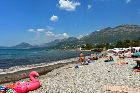 Słoneczny dzień na plaży w Barze, Czarnogóra. Ludzie opalają się, a w tle widać góry. Różowy flaming unosi się na kamienistej plaży.