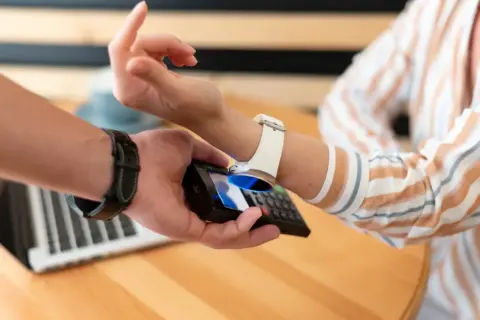 Zbliżenie na dłoń trzymającą telefon, który zbliża się do białego zegarka na ręce. Pokazuje, jak naładować zegarek telefonem.