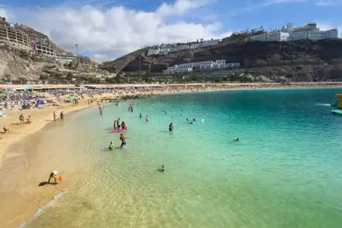 En livlig strand på Gran Canaria med solbadare, badande och en uppblåsbar vattenpark. En perfekt dag för att njuta av solen och havet.