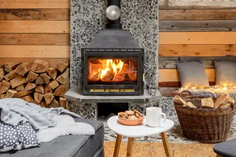 Przytulny kominek z palącym się drewnem. Obok stos drewna i kosz. Czy w kominku można palić węglem? Raczej nie, tu pali się drewnem.