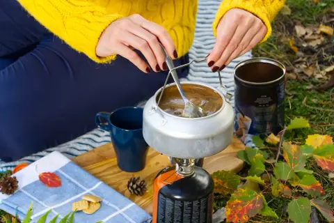 Preparando café en la naturaleza con recetas camping gas. Una tetera hierve sobre un hornillo, junto a una taza azul y galletas.
