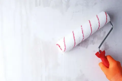 Ręka w pomarańczowej rękawicy wykonuje gruntowanie przed malowaniem, używając wałka z czerwonymi paskami.