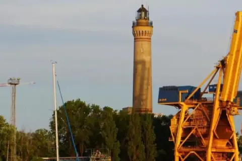Wysoka, żółta latarnia morska w Gąskach góruje nad portem, obok potężnych, żółtych dźwigów portowych i zielonych drzew.