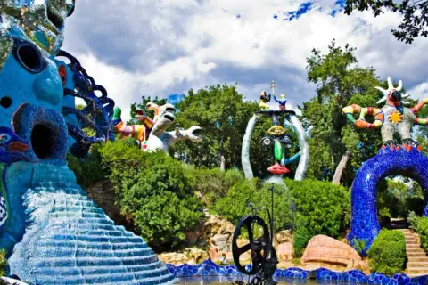 Sculture colorate e fantastiche popolano il **giardino dei tarocchi**, tra alberi verdi e cielo nuvoloso.