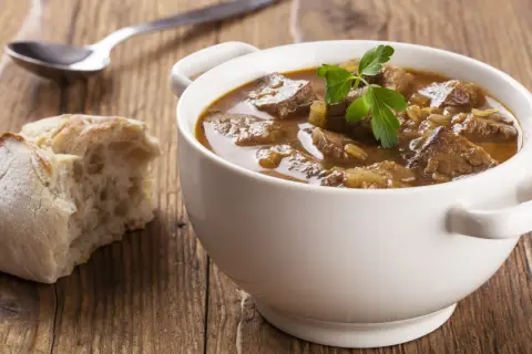 Aromaticzny, tradycyjny gulasz wołowy w białej miseczce, udekorowany natką pietruszki, obok kromka chleba i łyżka.