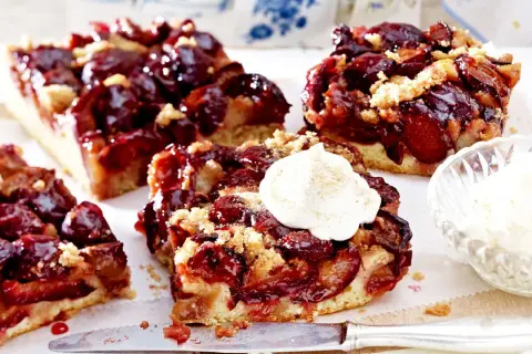 Saftige Pflaumenstücke auf einem lockeren Teig, bestreut mit Streuseln. Ein Stück gesunder Pflaumenkuchen mit einem Klecks Sahne.
