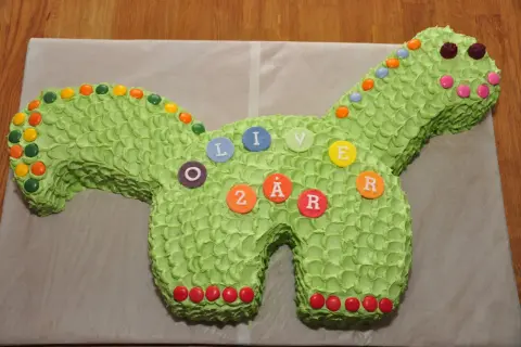 Enkel dinosaurietårta i ljusgrönt med godisdekorationer. Tårtan är formad som en dinosaurie och har bokstäver och siffror som bildar "OLIVER 2 ÅR".