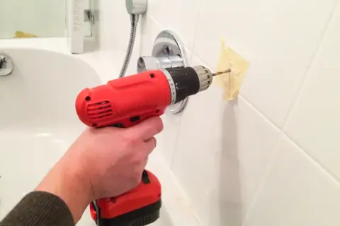 Une main tient une perceuse rouge pour percer du carrelage blanc, avec du ruban adhésif pour guider le foret.