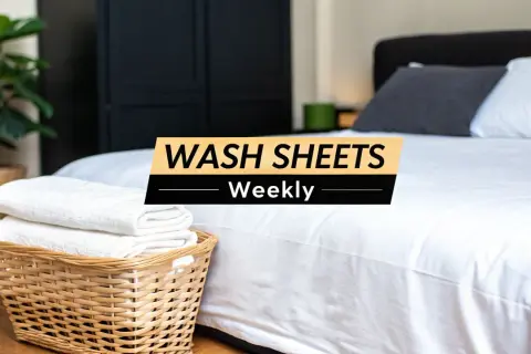 A clean bedroom with a neatly made bed. A basket of folded white towels sits beside the bed. Text overlay reads: "WASH SHEETS Weekly.