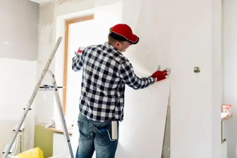 Mann mit roter Kappe und Handschuhen kleistert Vliestapete ein, um eine Wand zu tapezieren.