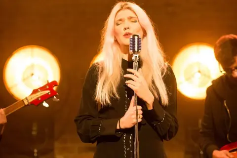 Wokalistka z blond włosami śpiewa przy mikrofonie, w tle zespół z gitarami i światła.