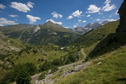Vue panoramique d'un col de tentes dans les Pyrénées, avec une route sinueuse traversant des prairies verdoyantes et des montagnes majestueuses sous un ciel bleu parsemé de nuages.