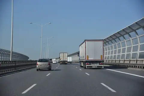 Jadę lewym pasem autostrady za ciężarówką. W oddali widać inne pojazdy i ekrany akustyczne.