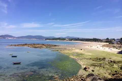 Vista de la playa de A Calzoa en Vigo, con gente disfrutando del sol y el mar. Barcas pequeñas flotan en aguas cristalinas.