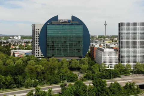Charakterystyczny budynek Radisson Blu we Frankfurcie, otoczony zielenią, obok nowoczesnych wieżowców.