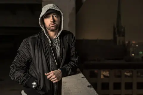 Eminem w czarnej kurtce bomberce i szarej bluzie z kapturem, oparty o betonową barierkę.