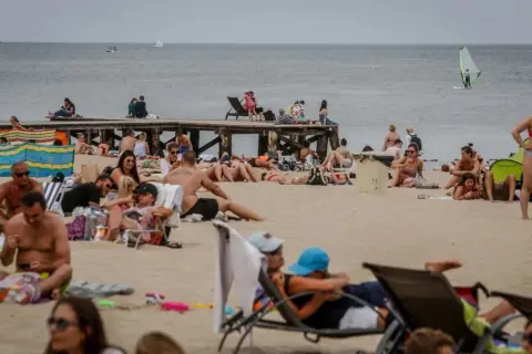 Tłum ludzi na plaży, opalanie się i relaks. Idealna pogoda na lipiec nad morzem, widać żaglówkę i wiatrak.