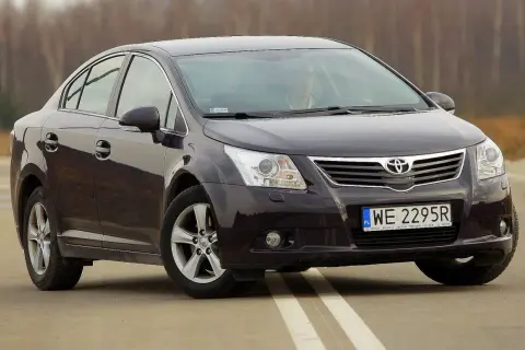Ciemny sedan Toyota Avensis na drodze. Kierowca za kierownicą. Szczegółowe dane techniczne tego modelu są dostępne online.