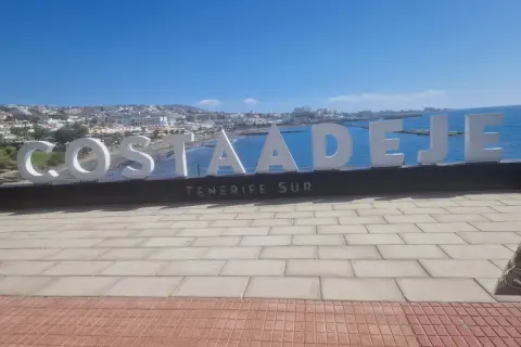 Napis "COSTA ADEJE TENERIFE SUR" na tle oceanu i miasta. Uważaj na słońce i tłumy, by w pełni cieszyć się wakacjami na Teneryfie.