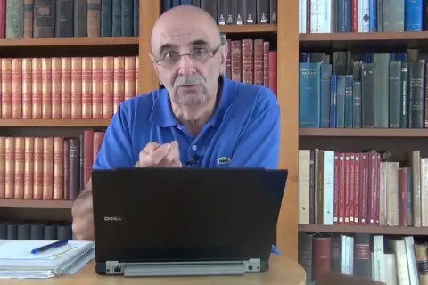 Un homme parle devant un ordinateur portable, entouré de livres. Il semble discuter de l'ad 07 état civil.