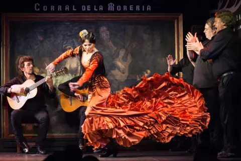 Una bailaora de flamenco, con un vestido naranja voluminoso, baila al son de guitarras. Es un espectáculo de flamenco vibrante.