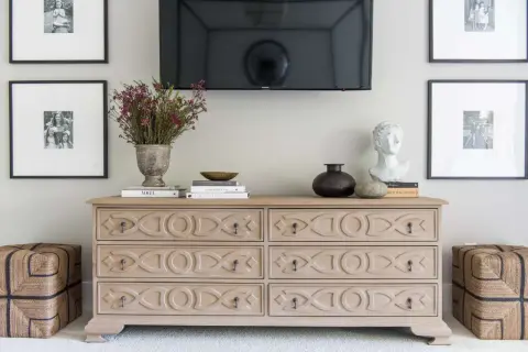 Ideas on how to decorate a dresser: a light wood dresser with decorative carvings, topped with books, a vase of flowers, a bust, and a dark vase.
