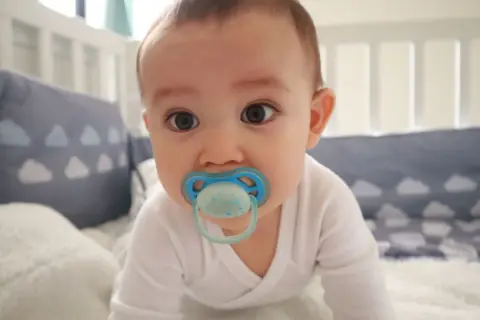A baby with a blue orthodontic pacifier in a crib. Are orthodontic pacifiers better for developing smiles?