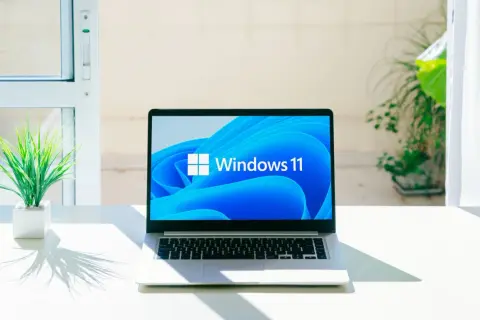 Laptop z logo Windows 11, gotowy na aktualizację 25H2. Na biurku obok rośliny, w jasnym pomieszczeniu.