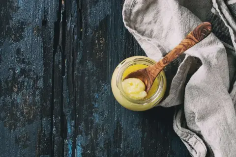 Ein Glas mit goldgelbem Ghee, daneben ein Holzlöffel mit einer Portion. Ghee ist geklärte Butter, ein vielseitiges Kochfett.