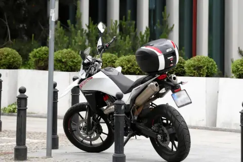 Biały motocykl z czarnym kufrem Givi zaparkowany na chodniku, obok słupków.
