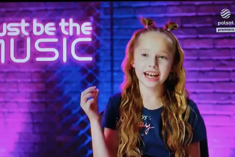 Lili chce śpiewać autor. Dziewczynka z burzą rudych loków i dwoma kucykami na głowie, na tle neonowego napisu "must be the MUSIC".