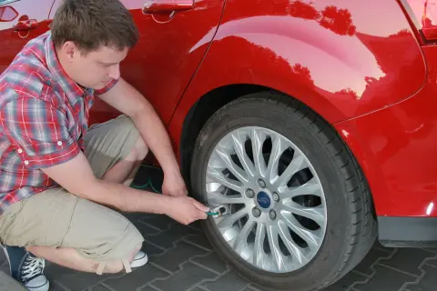 Mężczyzna w kratę sprawdza ciśnienie w kole czerwonego Forda. Wkrótce ruszy w dalszą podróż, tankując **benzynę**.