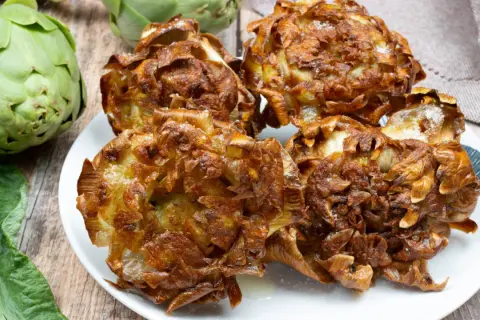 Piatto di carciofi alla giudia dorati e croccanti, con carciofi freschi sullo sfondo.