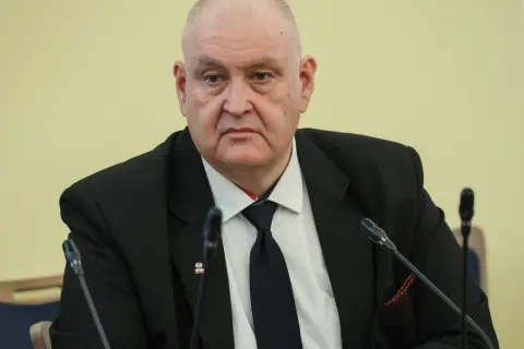 Prezes Trybunału Konstytucyjnego w garniturze, z przypinką z godłem Polski, siedzi przy stole z mikrofonami.